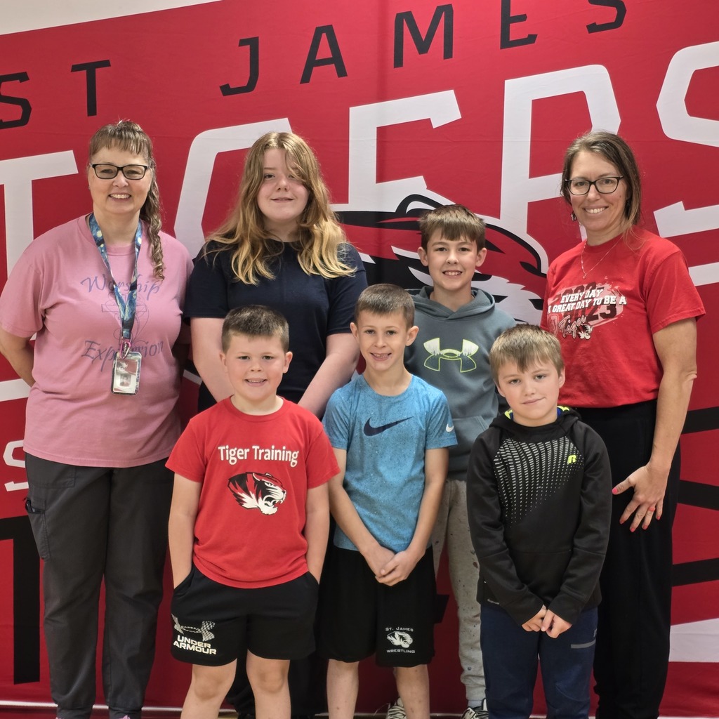 Five children and three adults stand together in front of a red wall with the word "James" on it.