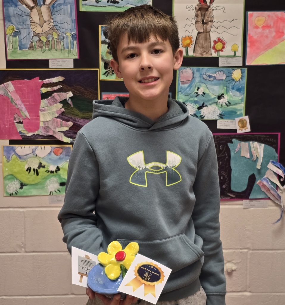 A boy in a gray hoodie stands in front of a wall with colorful artwork. He holds a trophy with a flower.