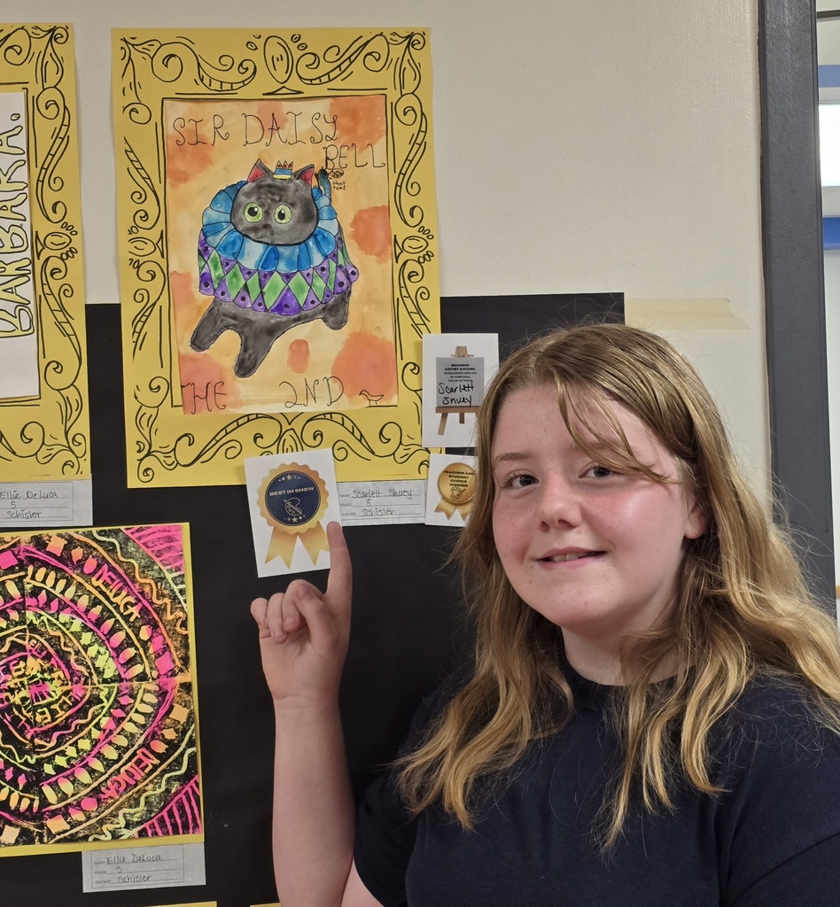 A girl with long blonde hair smiles at the camera. She points at a colorful cat art on the wall.