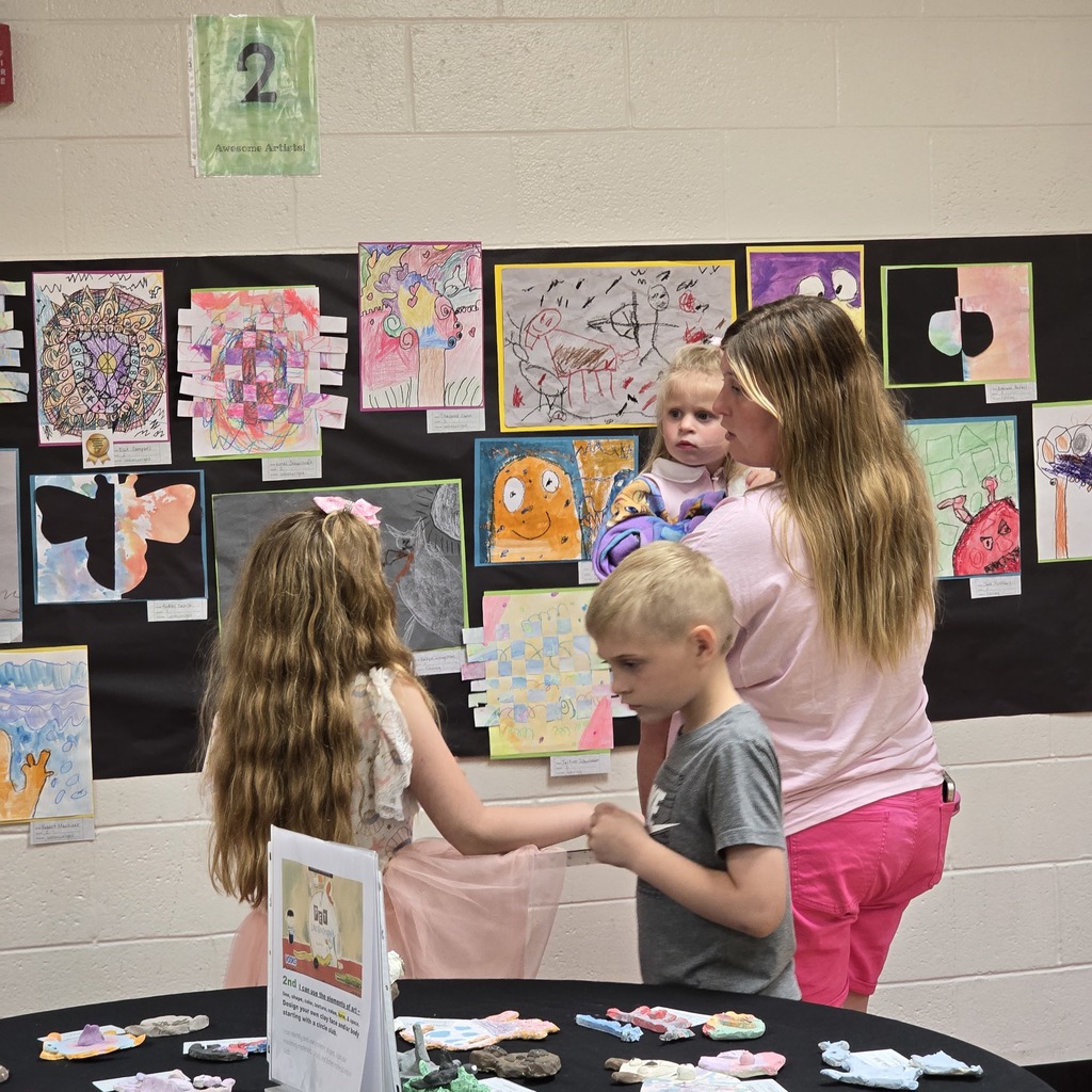 A room with artwork on the wall. A woman holds a baby and a child stands in front of a table.