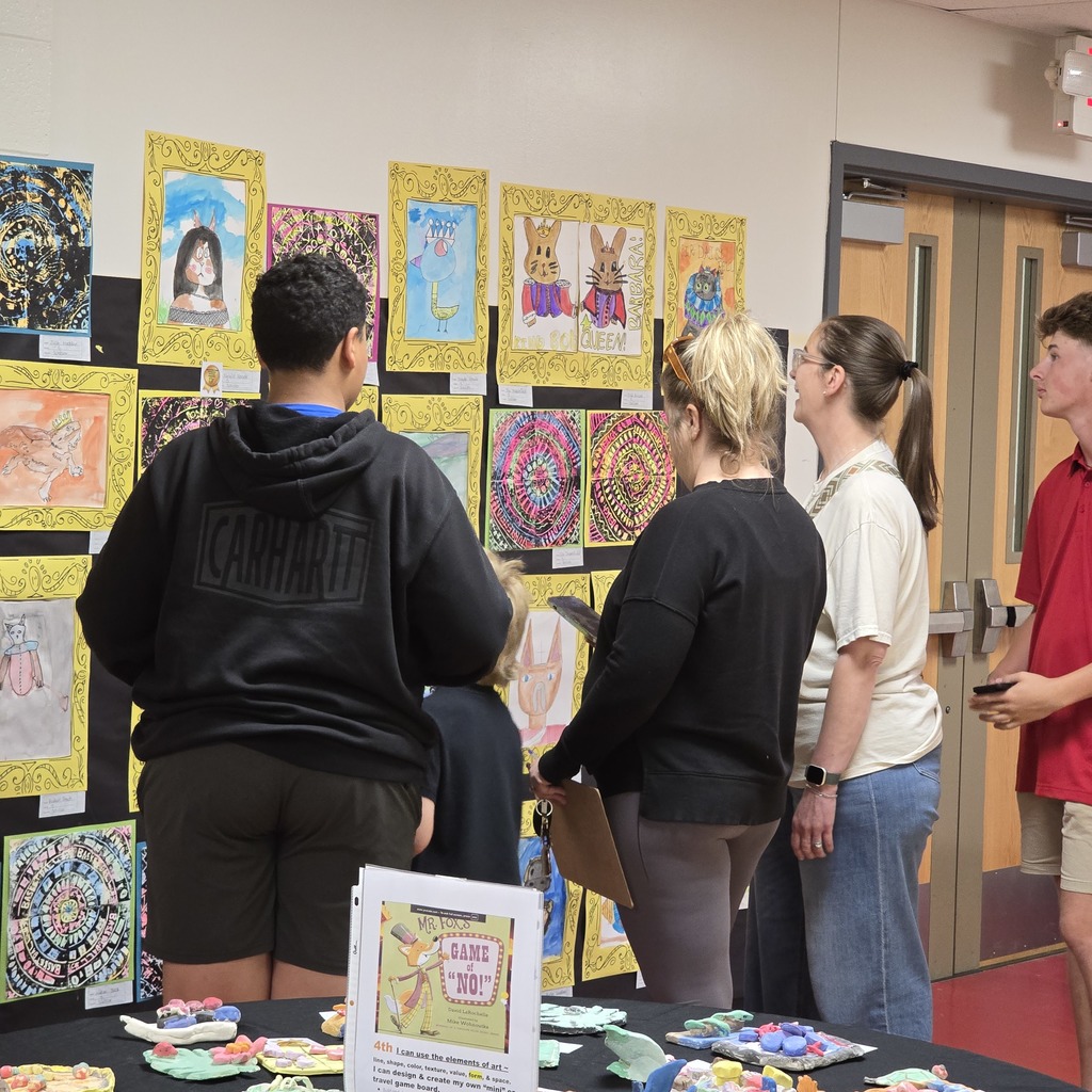 Several people view colorful artwork on a wall. A table holds paper crafts.