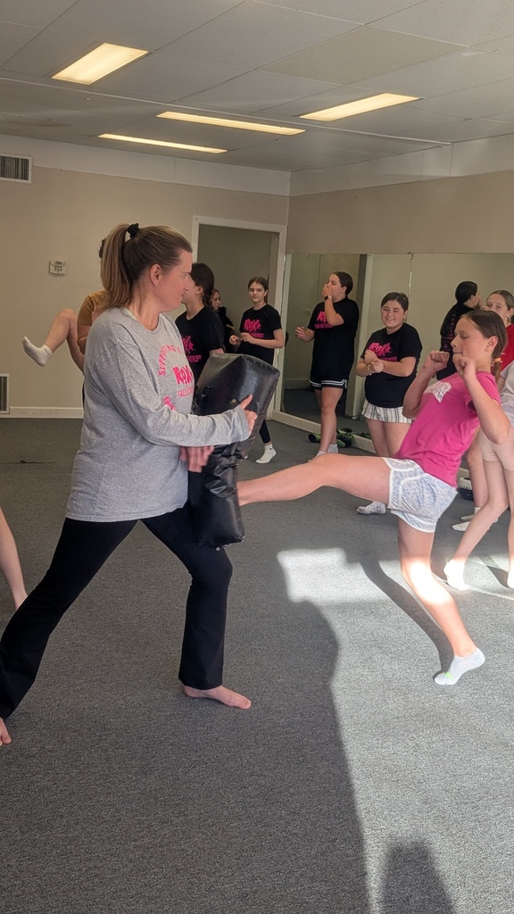 Multiple people in a room practicing martial arts with one woman demonstrating a kick to a padded bag.
