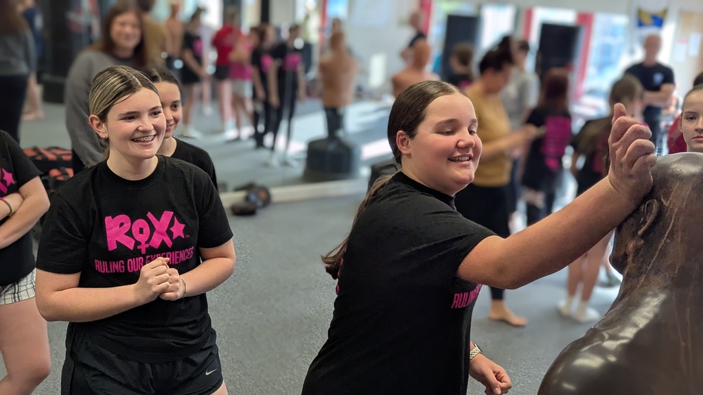 Women in black t-shirts, some smiling, participate in a fitness class. A woman leads by holding onto a punching bag.