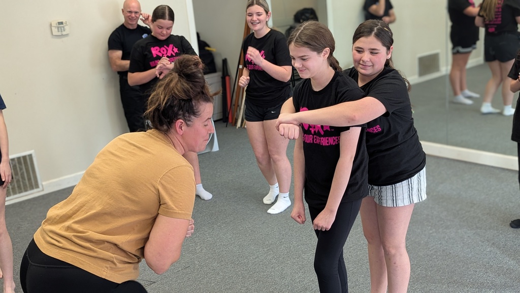 Group of young women in dance studio, wearing black t-shirts. One instructor in yellow shirt.