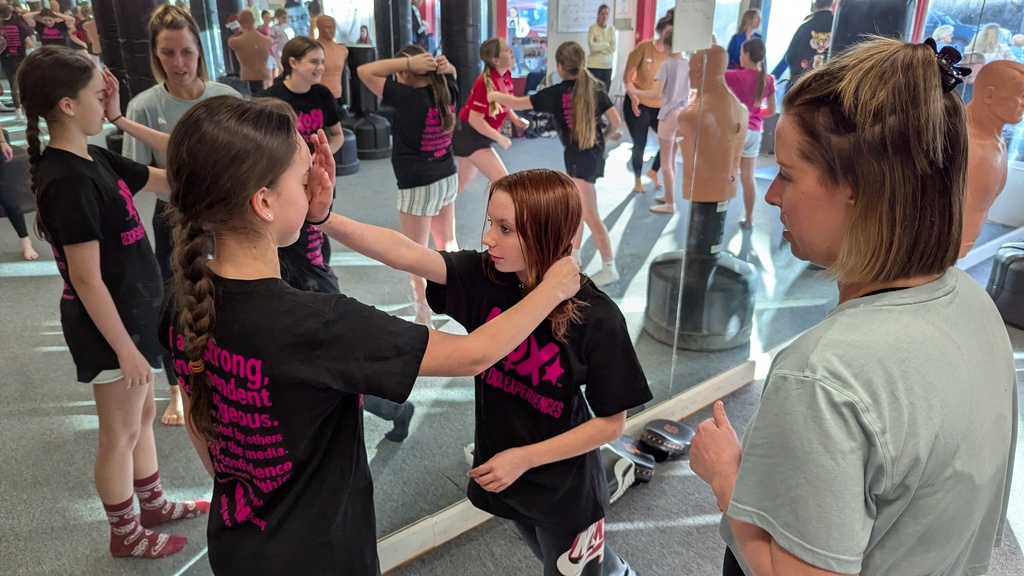 Two women in matching black T-shirts with pink writing and another woman in gray. They are in a mirrored room.