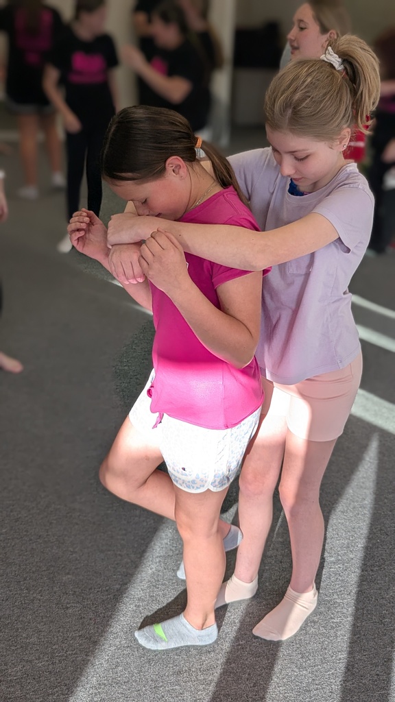Two girls in dance attire face each other, one bent over while the other holds her arms. Other girls stand behind them.