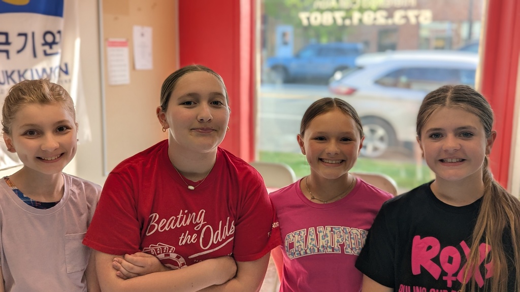 Four girls with smiling faces, wearing different colored T-shirts, standing close together. The background shows a glass window with a parked vehicle.