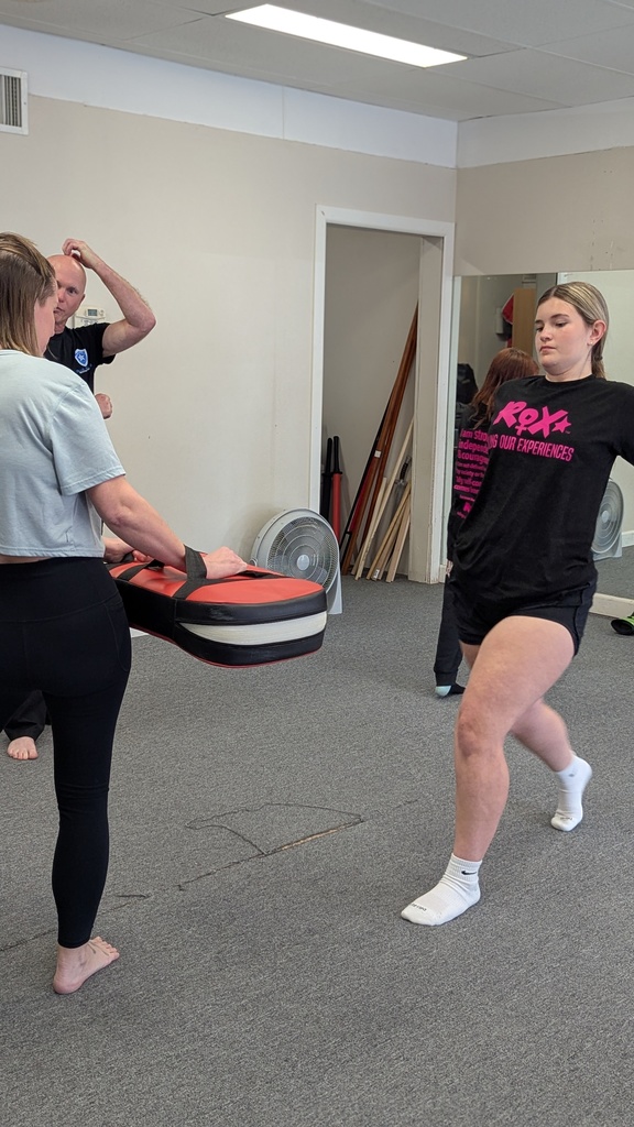 Three people in a room with exercise equipment and mirrors. A woman holds a training pad while another kicks.