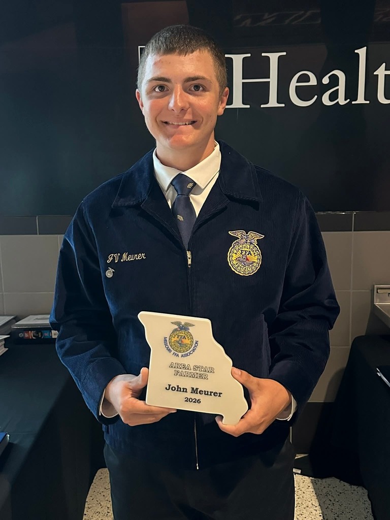 A person stands holding an award plaque in both hands. They wear a blue jacket and tie. Behind them, a wall displays "Health".