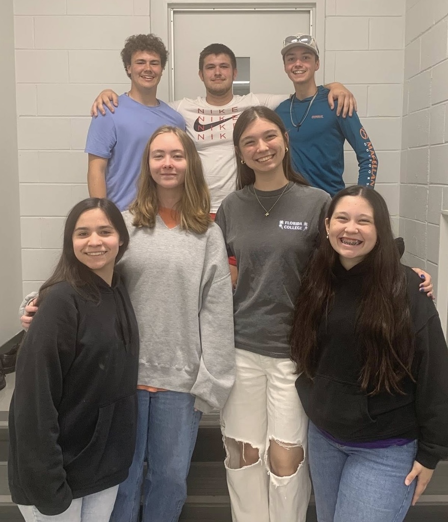 Six people in light-colored tops and pants stand on stairs, smiling, against a white wall.