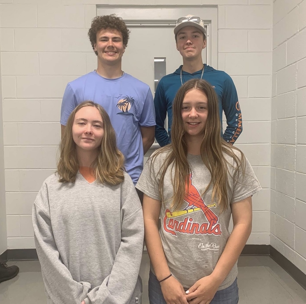 Four young people stand together in a hallway, smiling at the camera. Two wear blue shirts, one has a Cardinals shirt. White walls surround them.