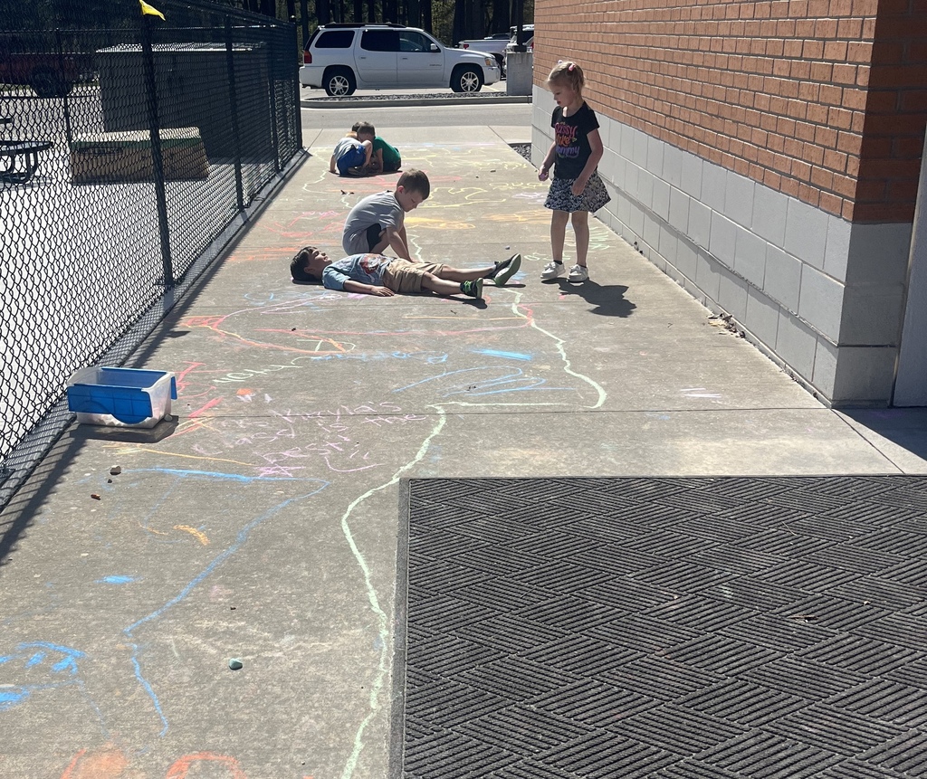 Four kids draw with chalk on a sidewalk. One lies on the ground. A brick wall and fence in background.