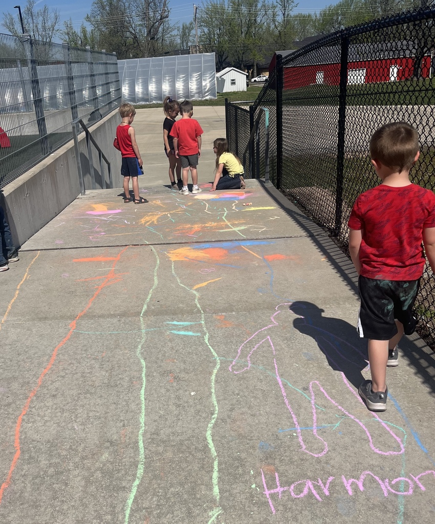 Children in red shirts draw chalk outlines on a concrete path. A gate and a fence are in the background.