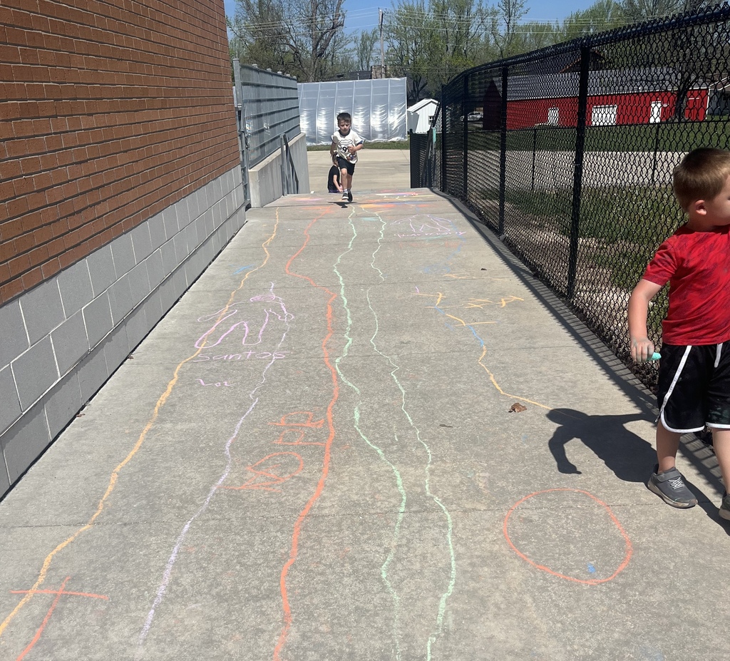 Two children run on a sidewalk with chalk lines. One child runs ahead, another follows. Background includes a brick wall, fence, and grassy area.