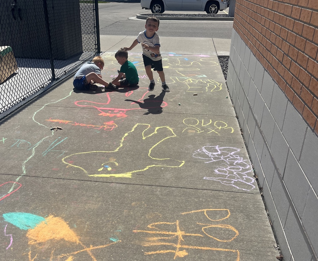 Three children draw on the sidewalk. One boy holds another's hand, while another sits. A brick wall and a fence are in the background.