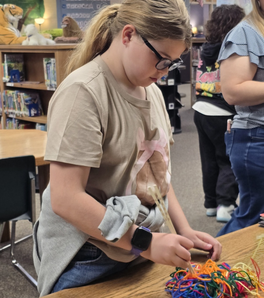 A young girl in a tan shirt and glasses works on a project with multicolored yarns at a table.