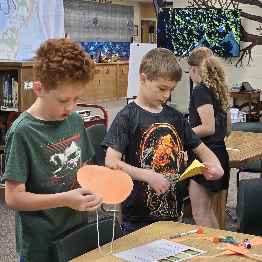 Two children are focused on making paper crafts at a table, with one holding a round orange paper.