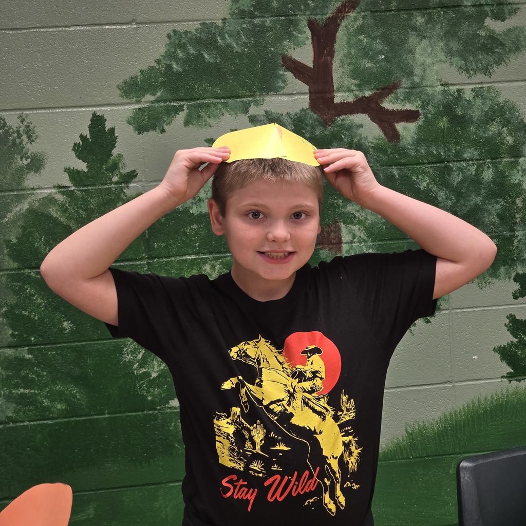 A young boy in a black T-shirt smiles while holding a yellow paper over his head.