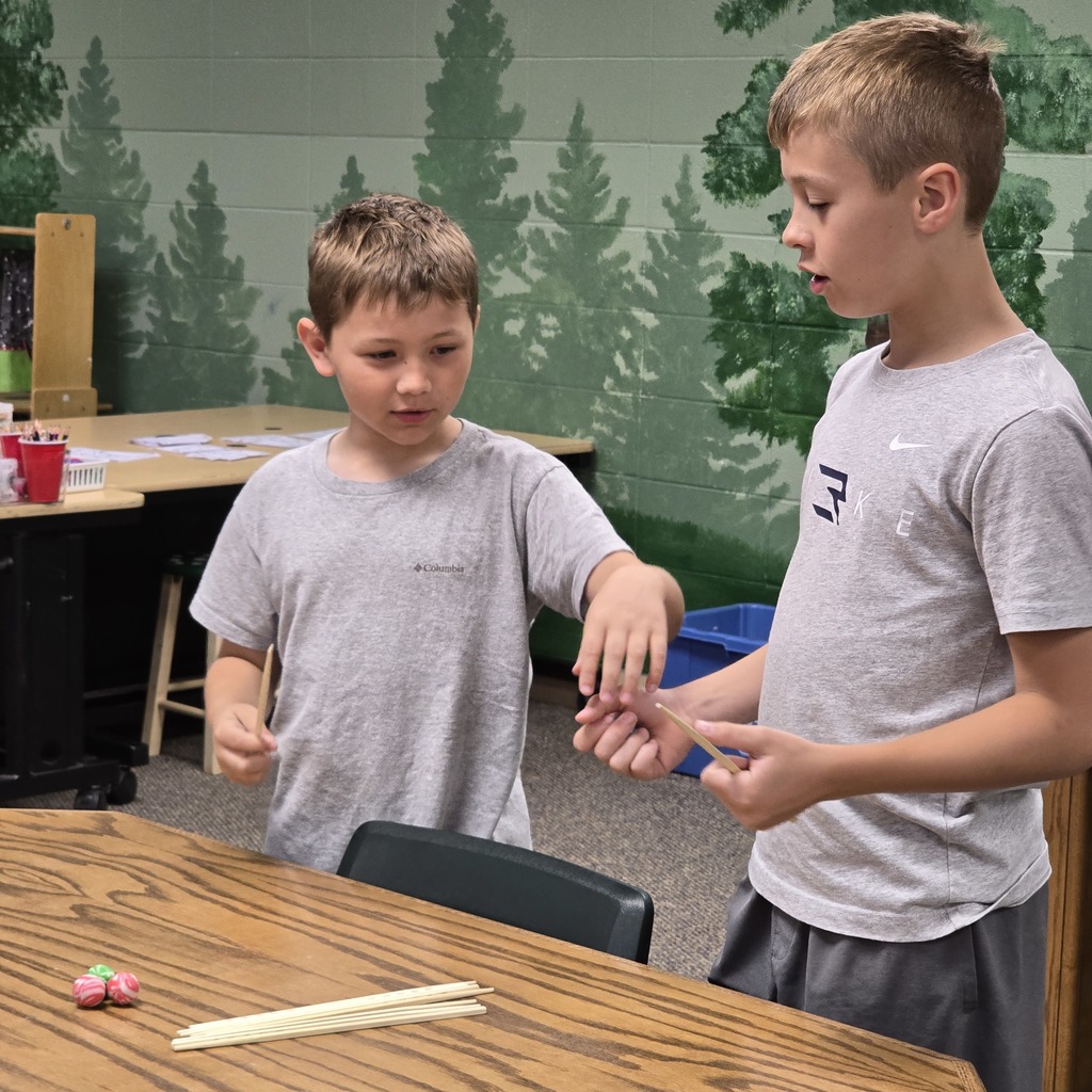 Two boys in gray shirts and pants in a room with trees on the wall, one holding sticks.
