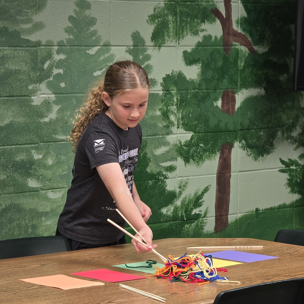 A girl manipulates colored yarn with chopsticks on a table, with a painted forest backdrop.