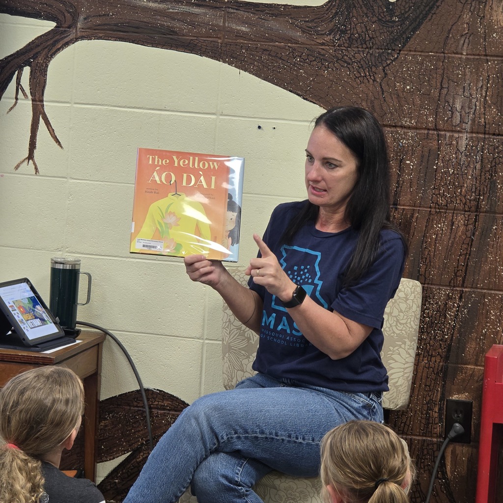 Woman in a blue shirt reading a book to a group of children sitting in a classroom.