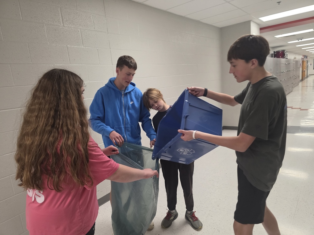 Four young people are carrying a blue box in a hallway, two of them wearing blue jackets.