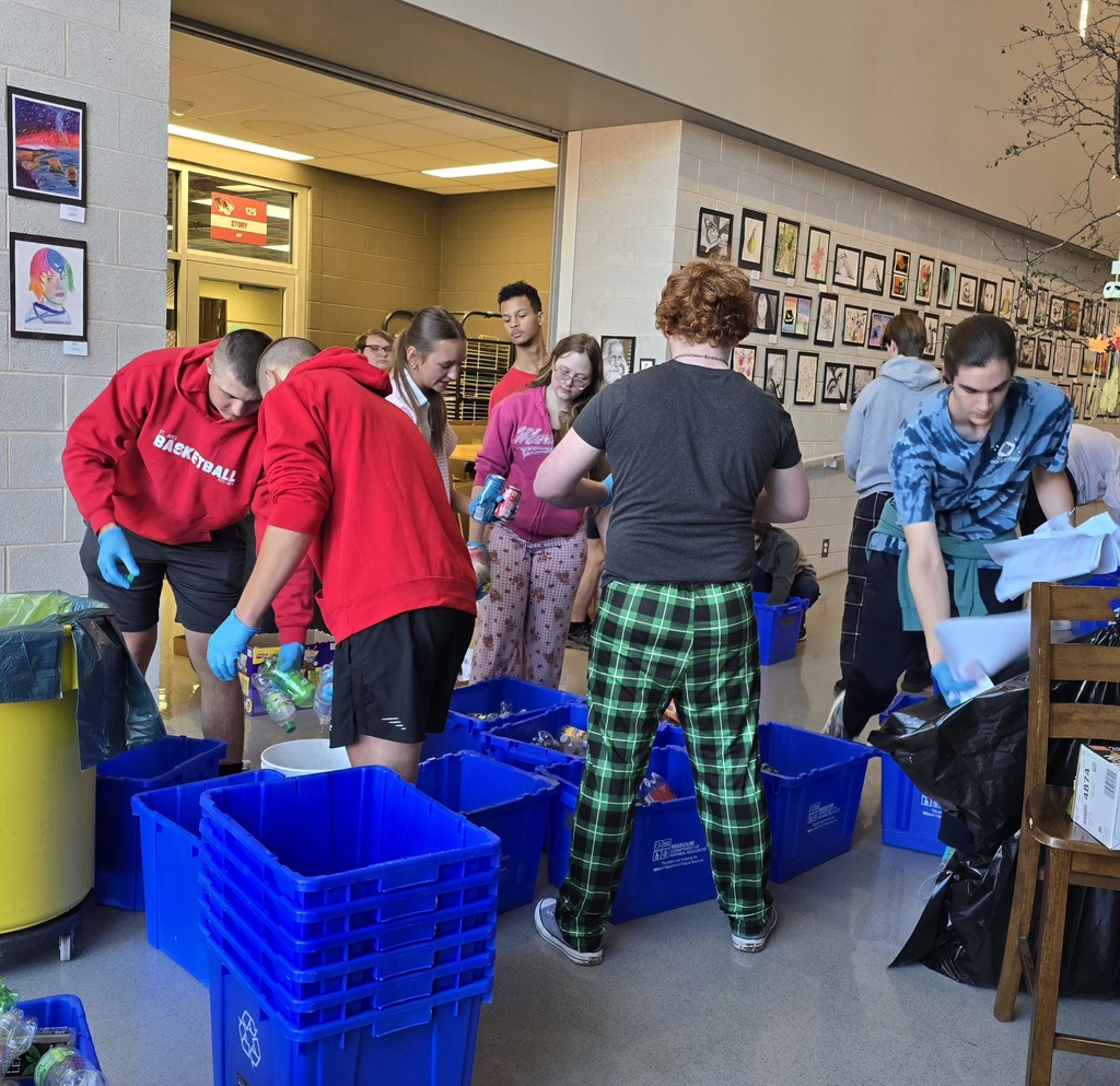 People sorting items into blue bins in a room with a yellow trash can, a chair, and artwork on the walls.