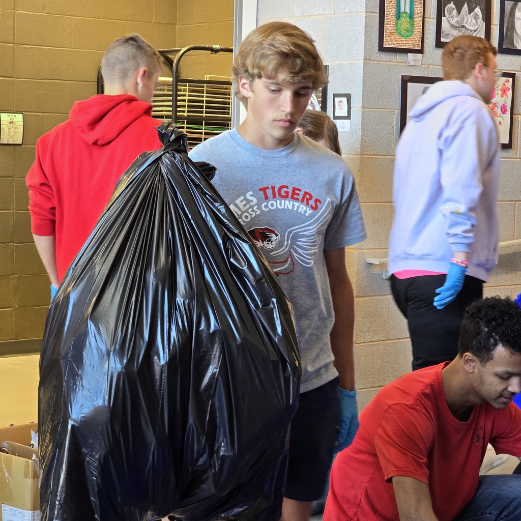 Four men are in a room with trash bags; one holds a bag, another wears a Tigers shirt, and two wear red shirts.