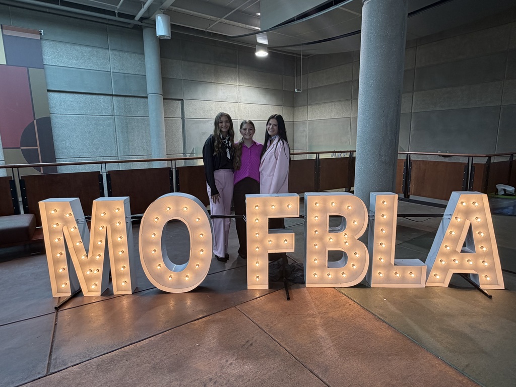 Three people stand next to a large light display spelling out "MOFBA" in a spacious room.