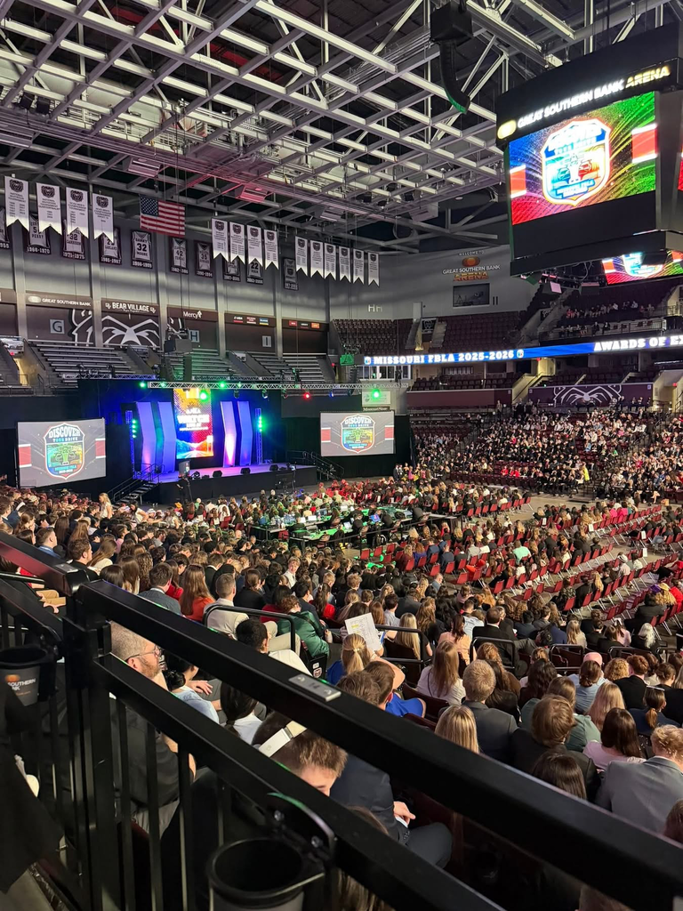An indoor stadium with many people seated in rows. Screens on the stage display logos. Flags hang from the ceiling.