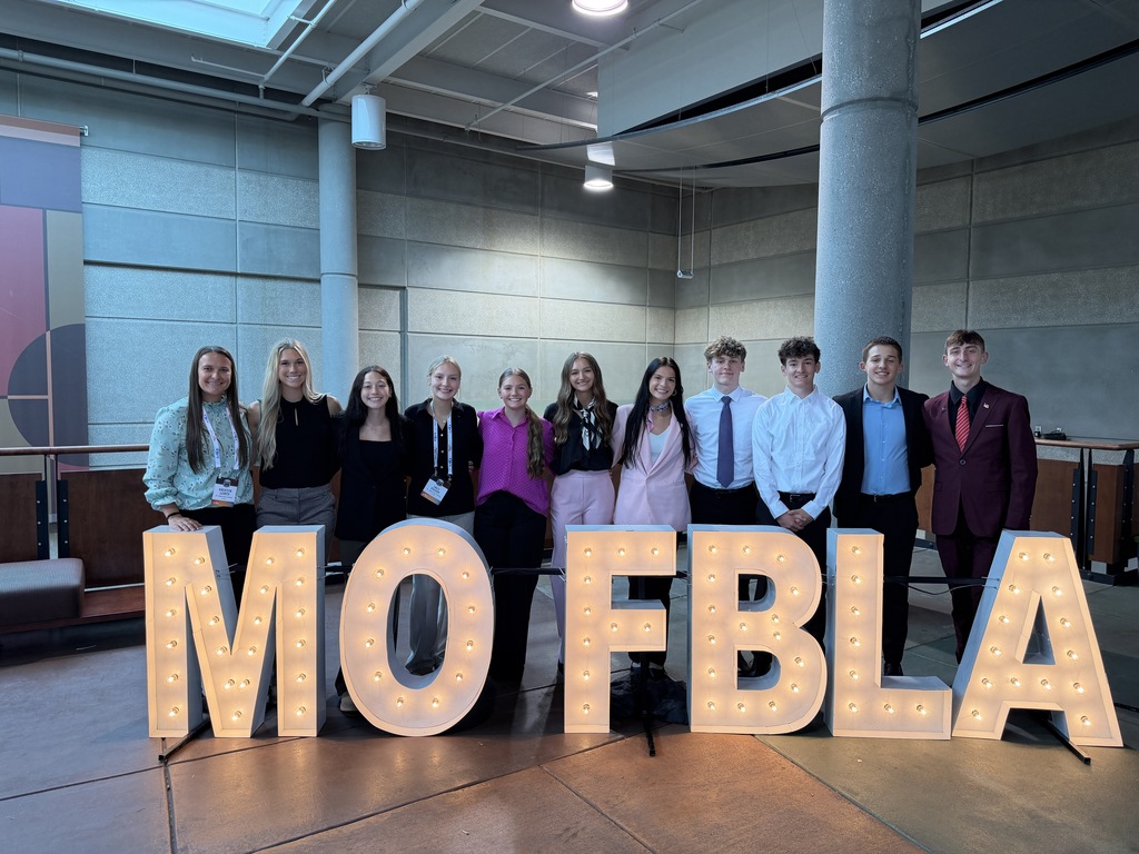 A group of students stands in front of large, lighted letters spelling "MOFBLAN" in an indoor room.