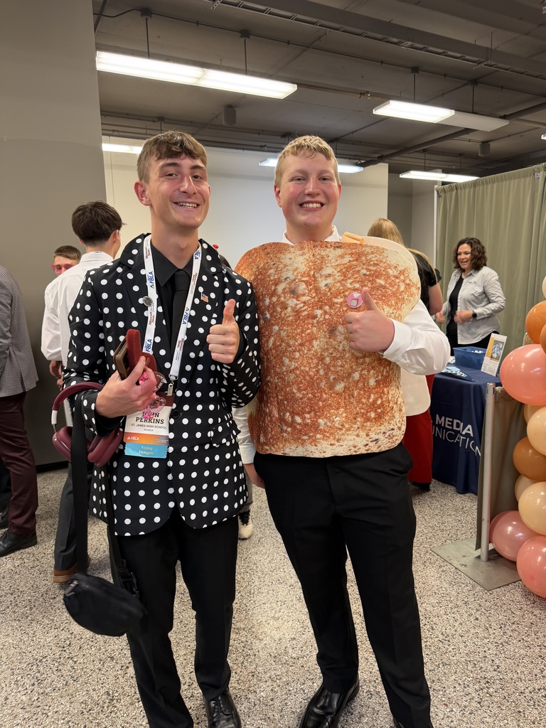 Two men stand close, smiling, one with a polka-dot shirt, the other wearing a toast costume. Behind them, people and balloons are in a room with white lights.