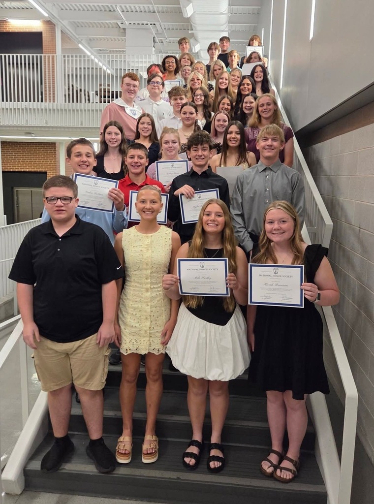 A group of students stand on stairs, holding certificates. They wear casual summer clothing. Behind them, there are railings and a hallway.