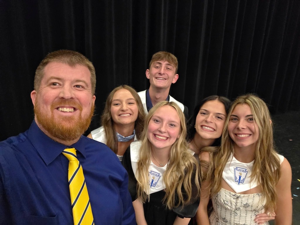 A group of people, including a man and five women, are smiling and standing together in front of a black backdrop.
