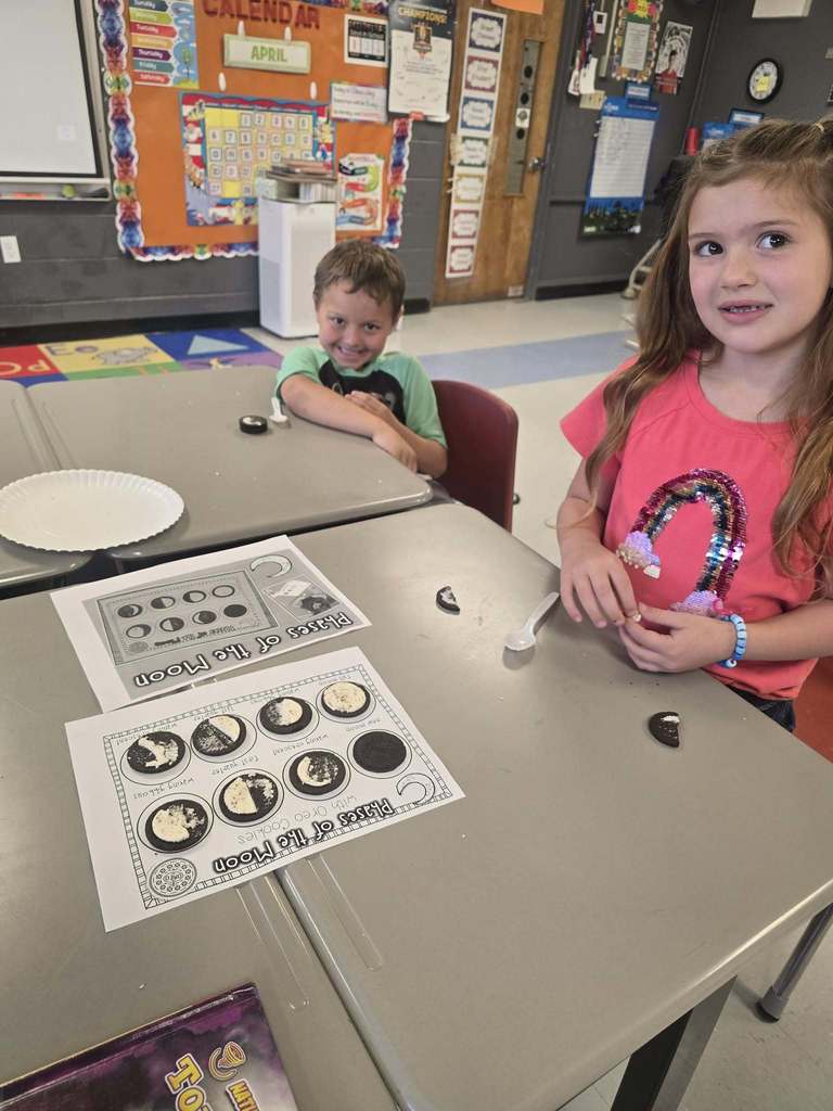 Two children seated at a table, engaged in an activity with various objects and papers.