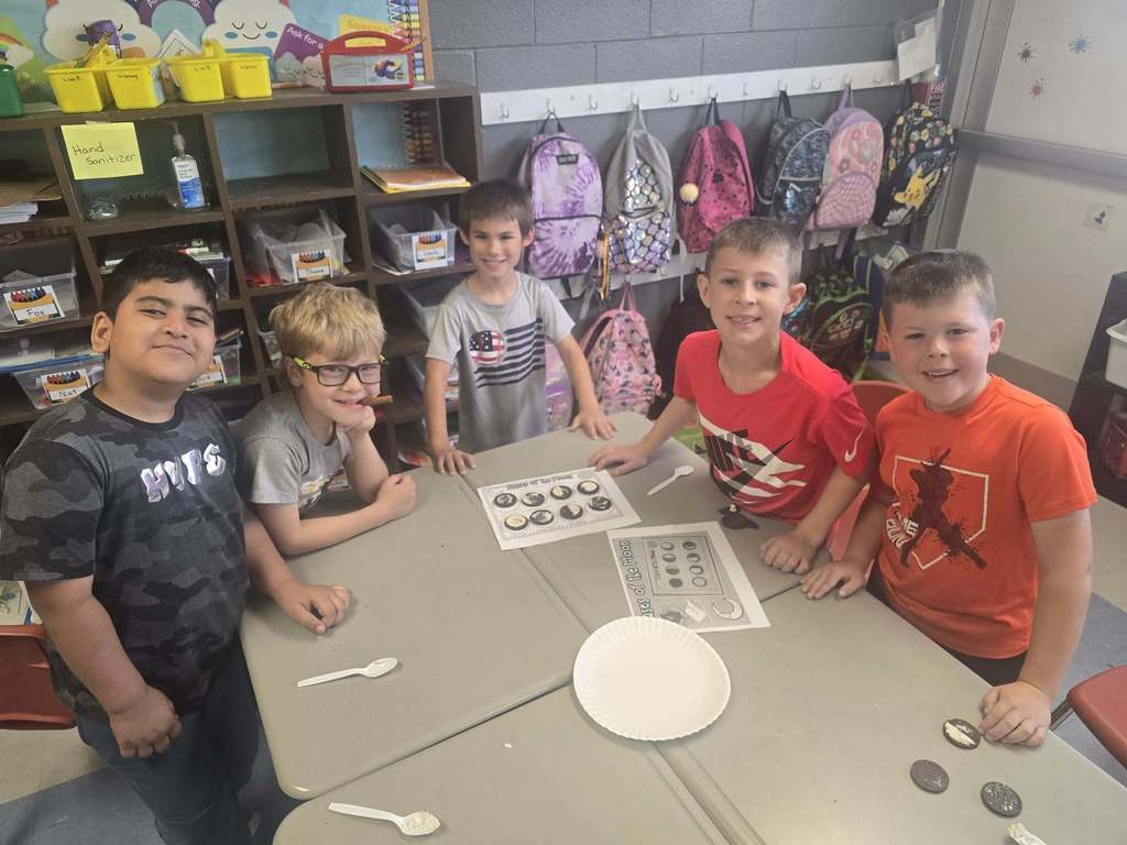 Five boys at a table, holding spoons and a plate, with a bag of coins.