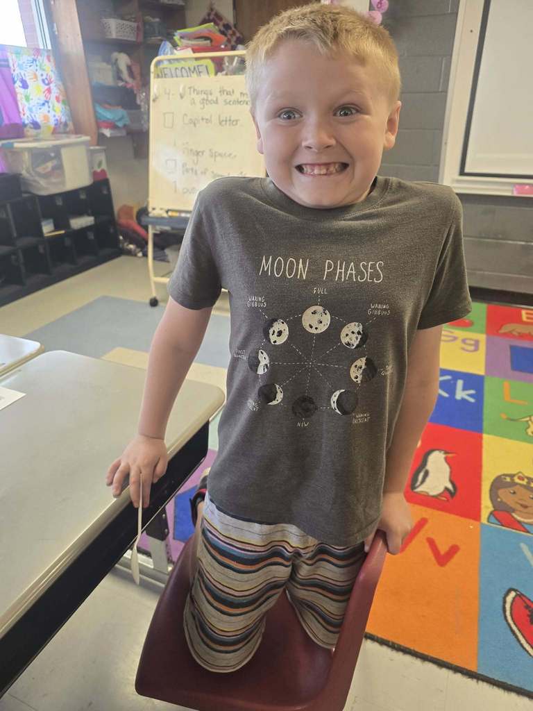 A child stands in a classroom, wearing a gray T-shirt with moon phases. He smiles, standing on a chair.