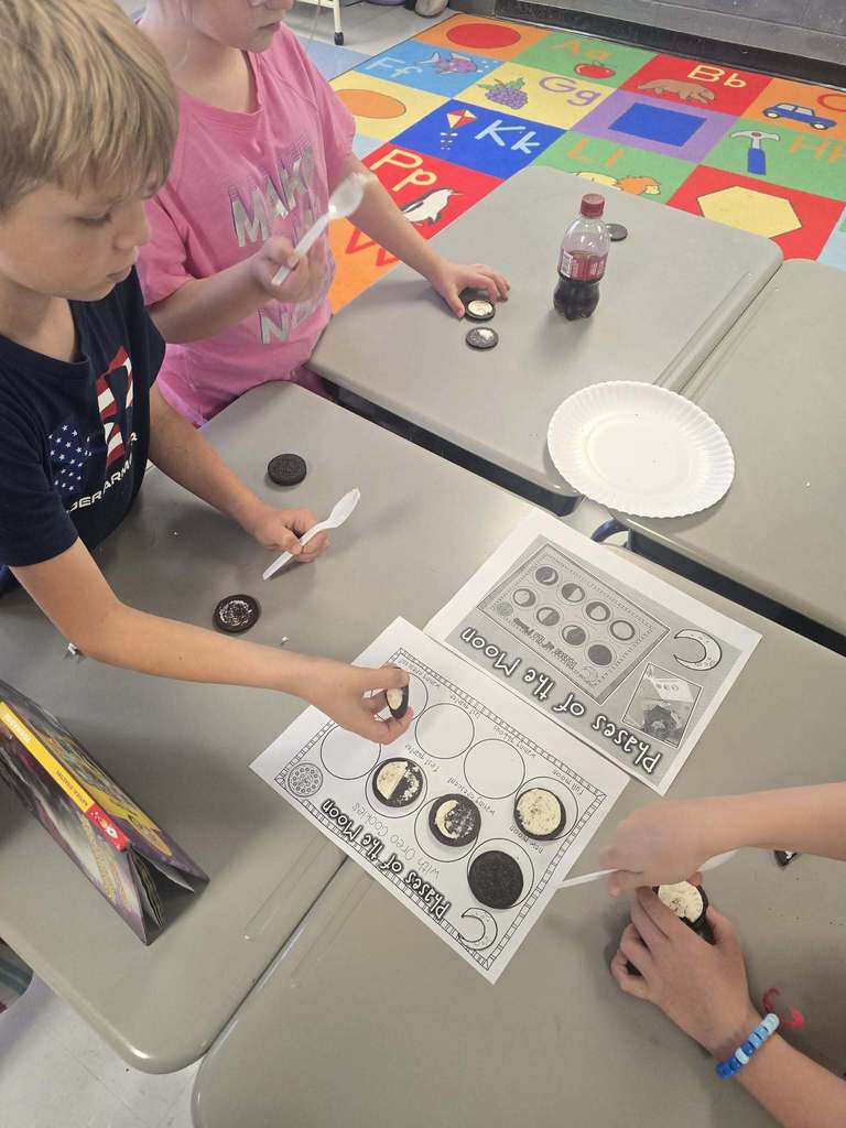 Children seated at a table, working on a project with cookies and a paper guide, surrounded by colorful floor tiles.