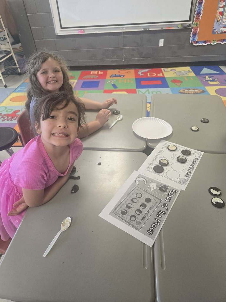 Two young girls sit at a desk, engaged in an activity with coins, paper, and spoons. Behind them is a colorful mat and a whiteboard.