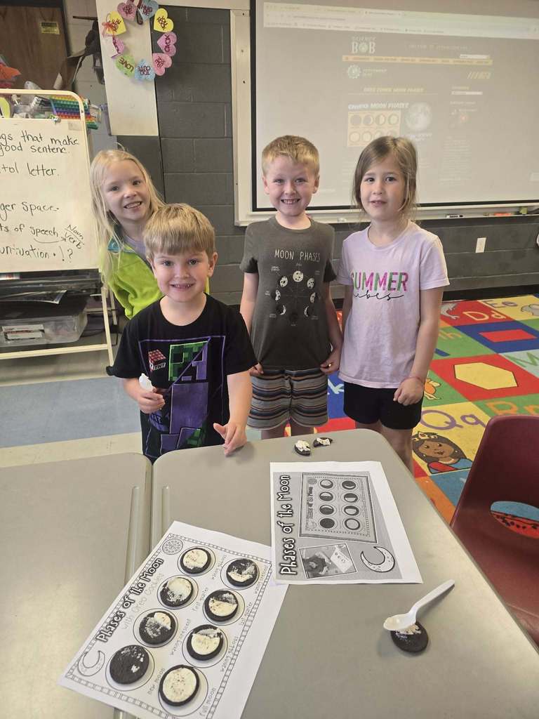 Four children in a classroom stand around a table with cookies and a worksheet on it.