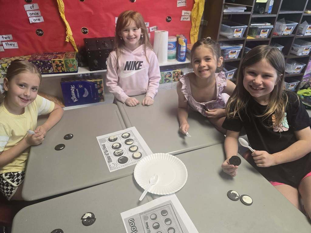 Four girls seated at a table with coins and a paper plate, engaged in a learning activity.