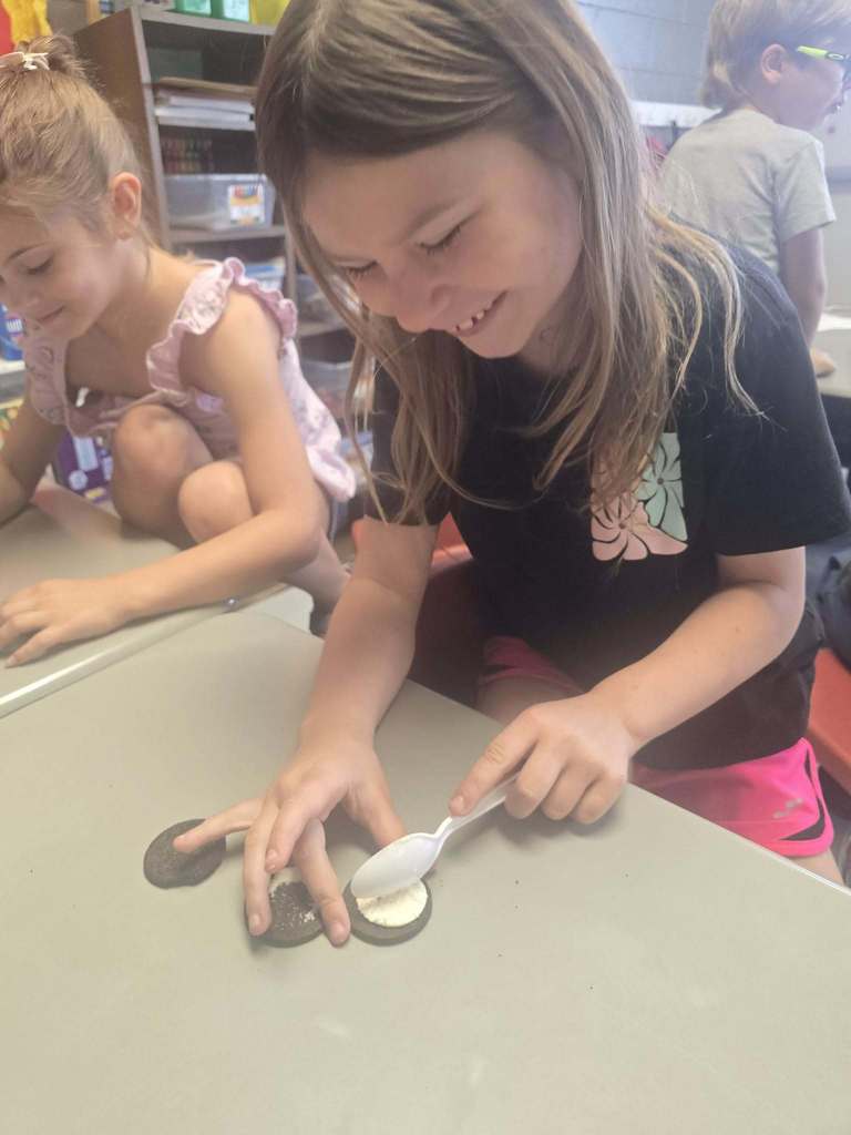 Two girls in a classroom setting, one playing with spoons and cookies on a table.