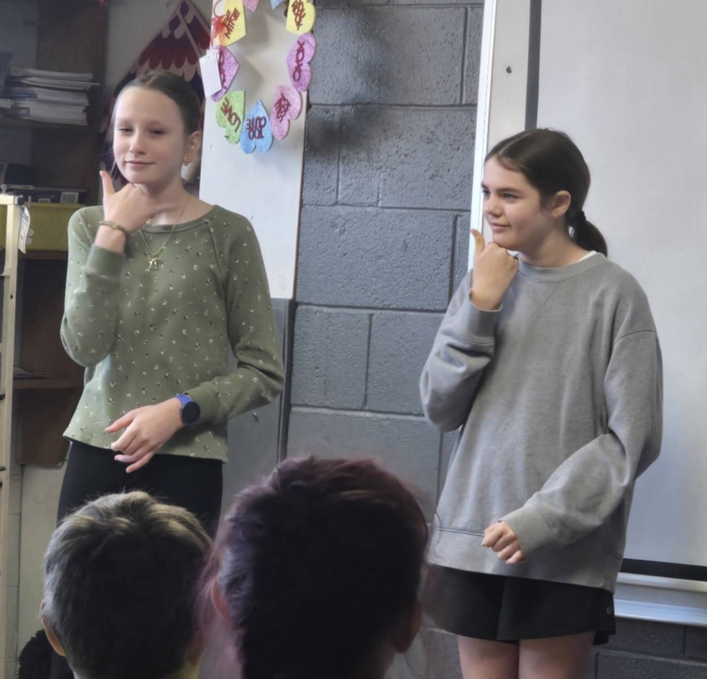 Two girls stand in a classroom, one in green and the other in gray. They face each other while speaking.
