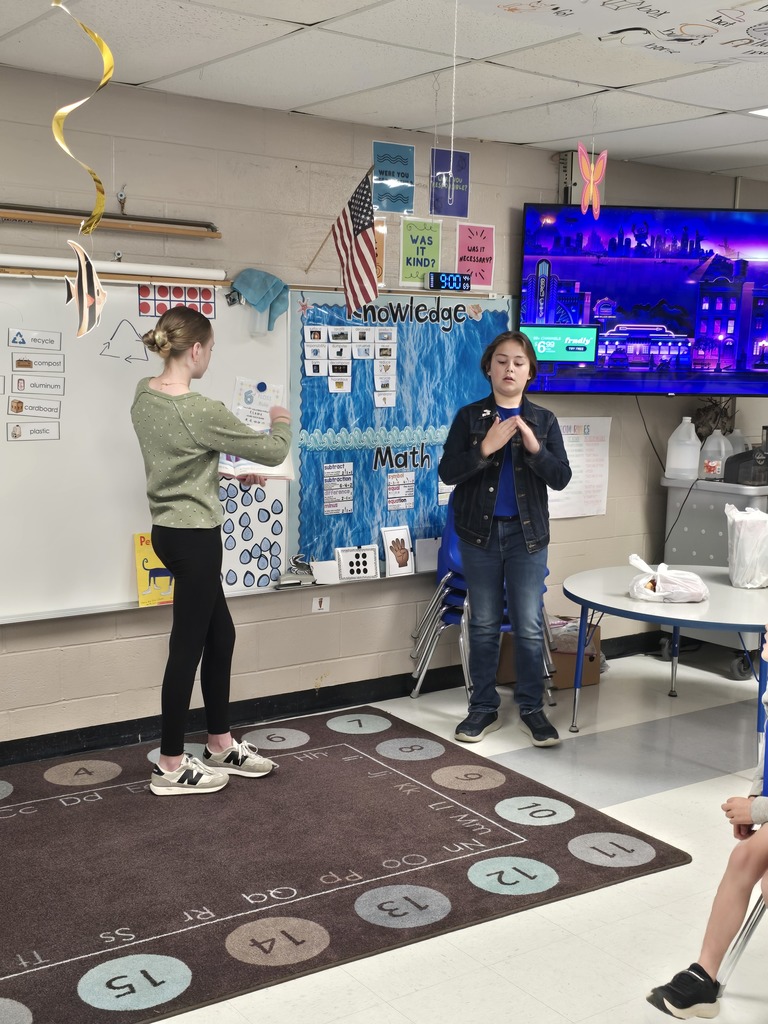 Two people stand in a classroom, one with hands clasped. A carpet with numbers and letters is on the floor.
