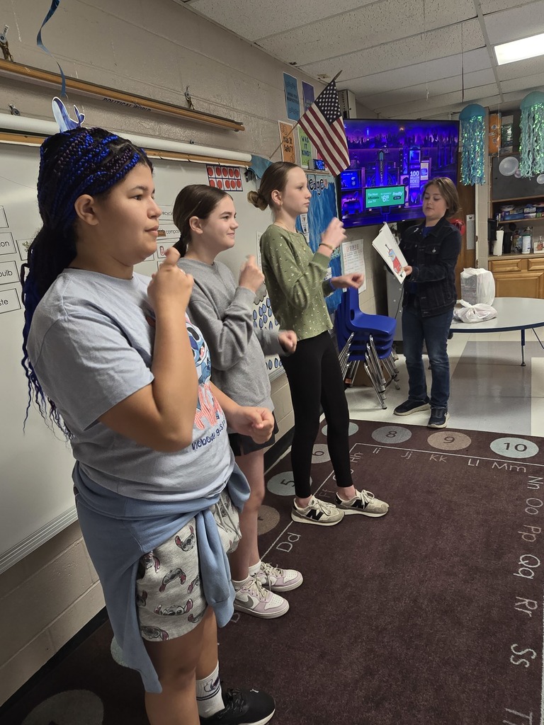 Four young women stand in a classroom. One holds a paper, another speaks, and the background shows a room with a rug, a table, and a television.