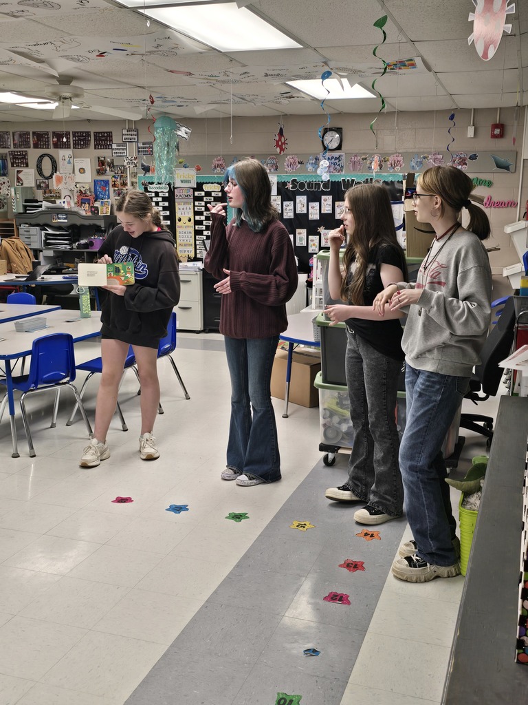 Four people stand in a room with colorful dots on the floor. Two hold books, one wears a gray sweatshirt, and another wears a maroon sweater.