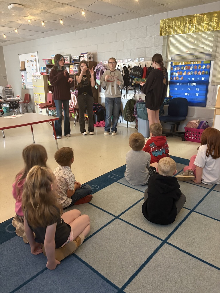 Several young children are seated on a blue carpet in a classroom, listening to adults standing and gesturing.