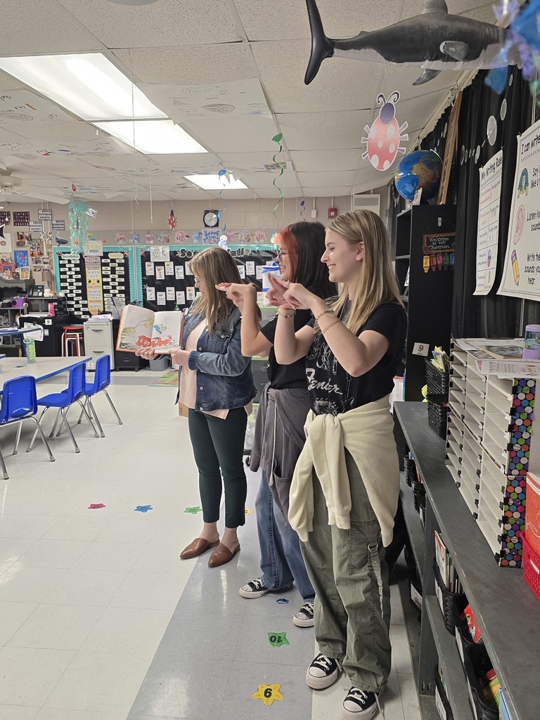 Three women stand in a classroom; one reads a book, and two gesture with hands. White walls and ceiling, tables, chairs, and decorative items.