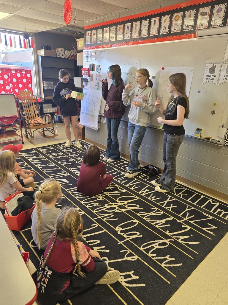 A group of students stands and sits on a carpet with alphabet letters, facing a whiteboard in a classroom.