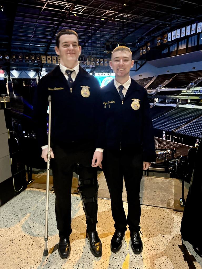Two men in matching navy uniforms, one using a cane, stand together in an empty stadium.